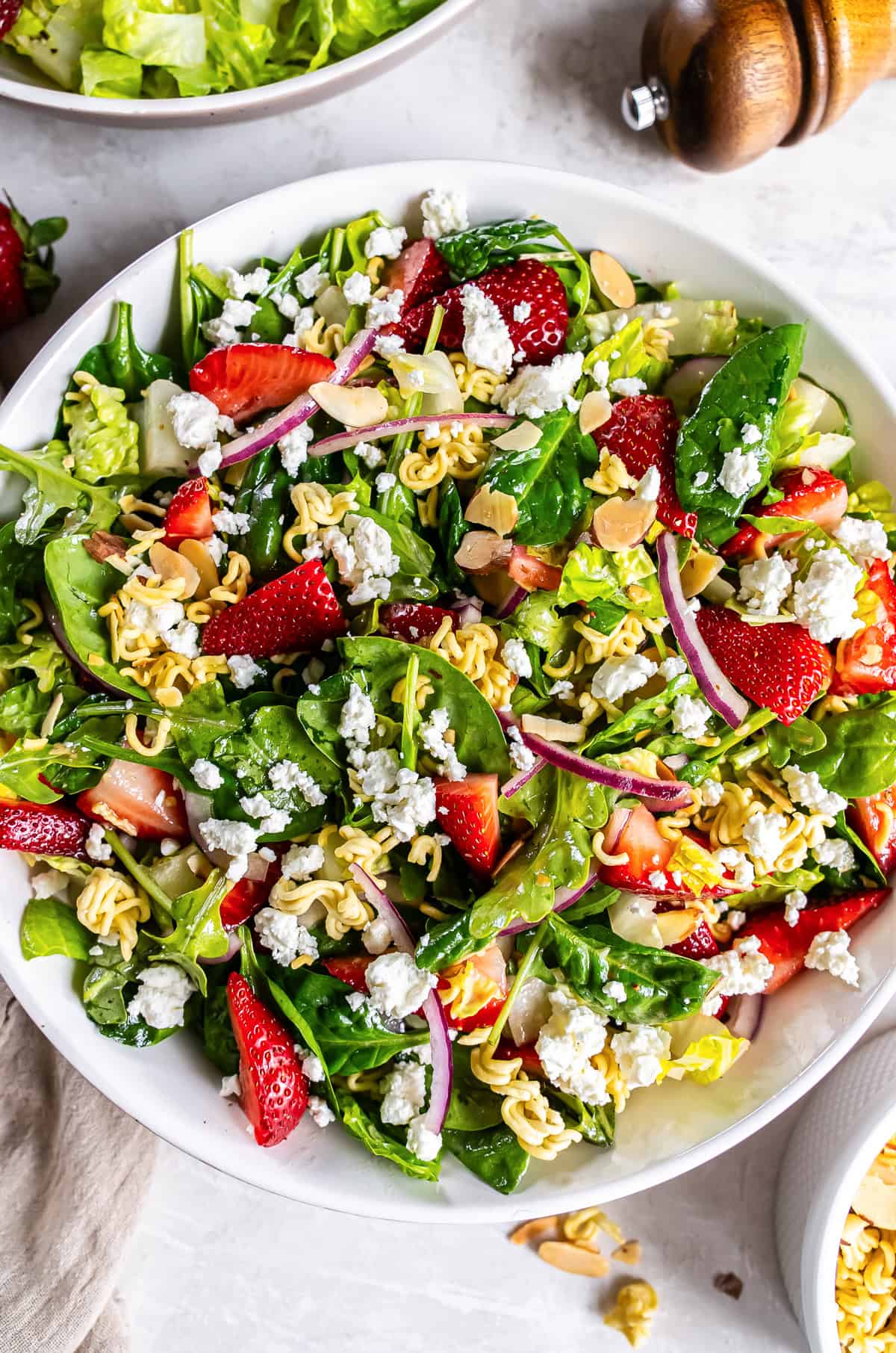 A salad with greens, strawberries, ramen noodles, and goat cheese in a large round white bowl.