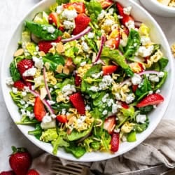A salad with greens, strawberries, ramen noodles, and goat cheese in a large round white bowl.