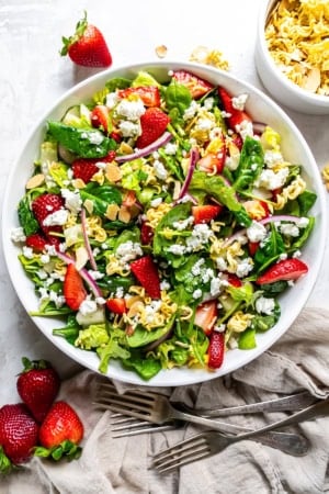 A salad with greens, strawberries, ramen noodles, and goat cheese in a large round white bowl.