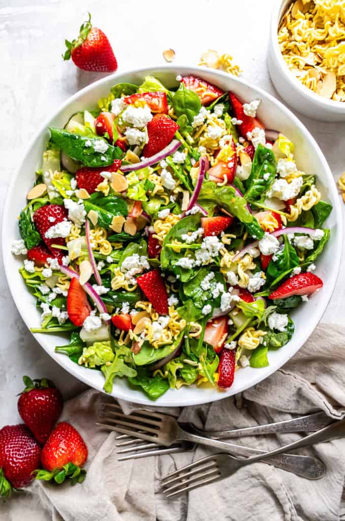 A salad with greens, strawberries, ramen noodles, and goat cheese in a large round white bowl.