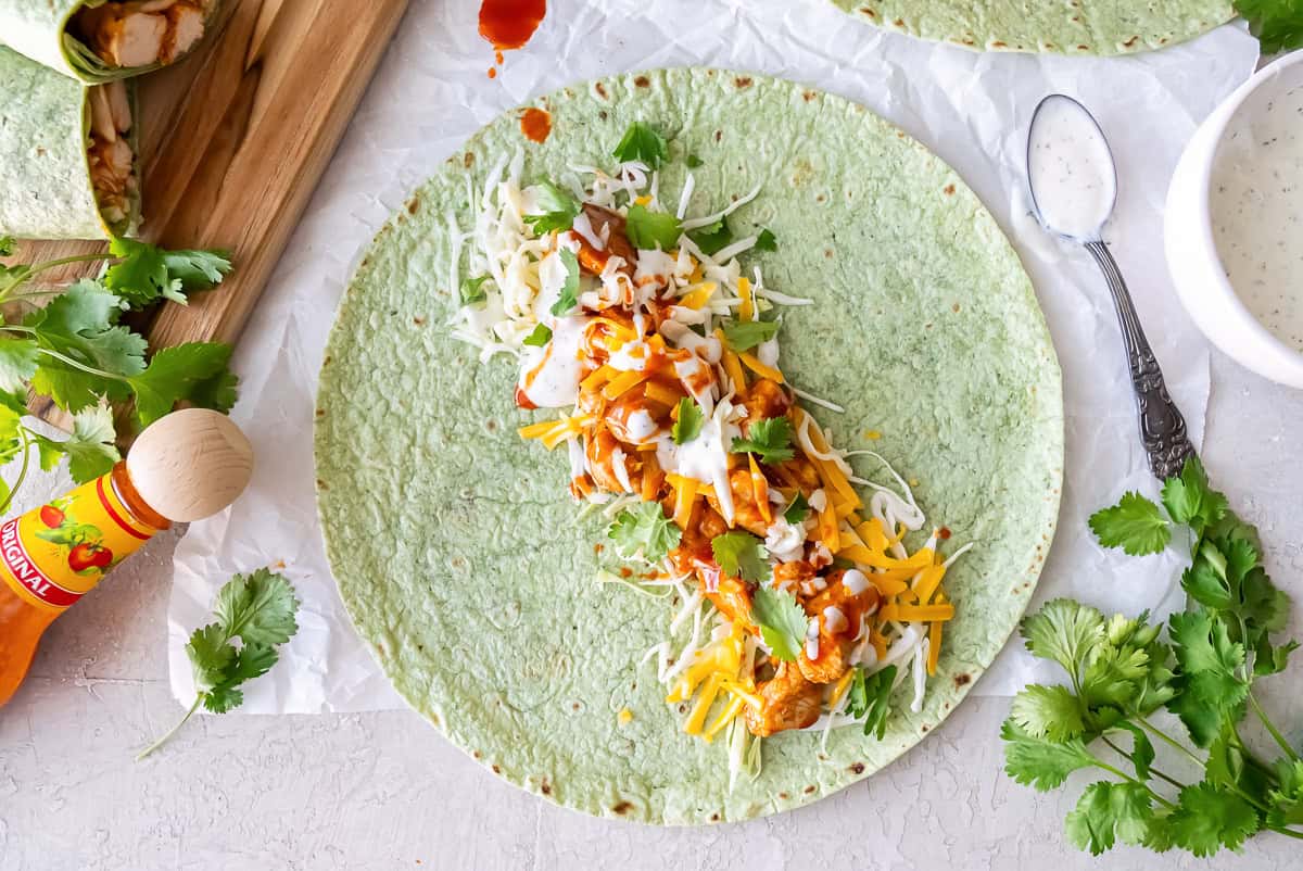 An open face bbq chicken wrap on a spinach tortilla next to a bottle of Cholula hot sauce and small bowl of Ranch dressing.