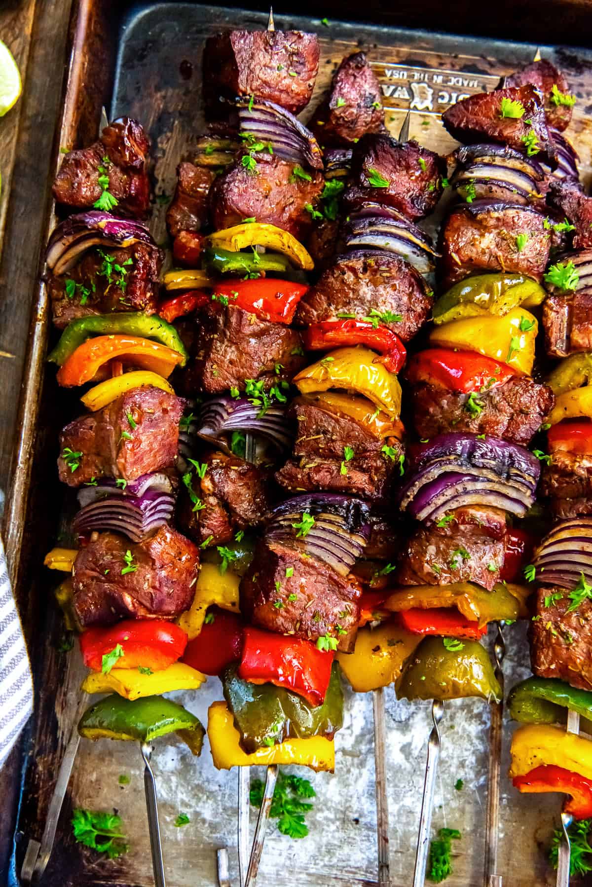 Kabobs with grilled beef, peppers and onions piled on a baking sheet.