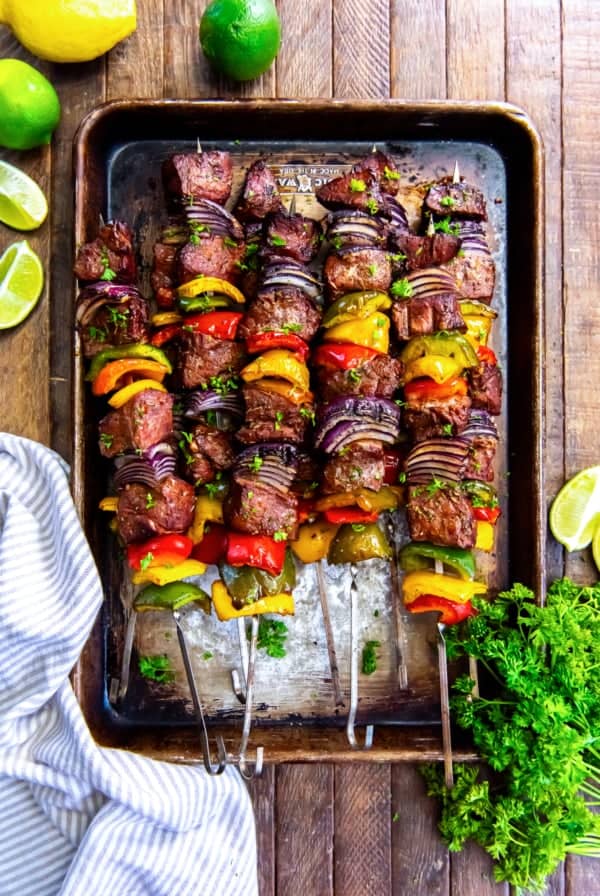 Grilled steak kabobs with peppers and onions on a baking sheet surrounded my lemons and limes.