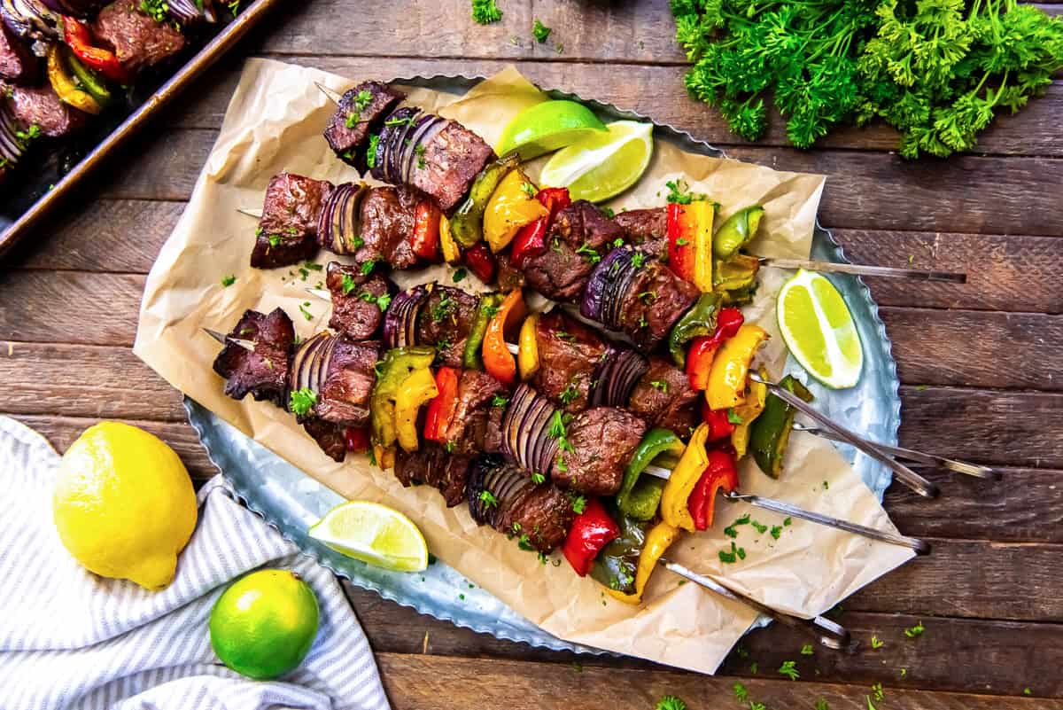Grilled steak kabobs piled on a platter lined with parchment paper surrounded by limes and lemons.