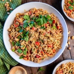 Peanut noodles with chicken in a large white bowl next to a small bowl of sesame seeds.