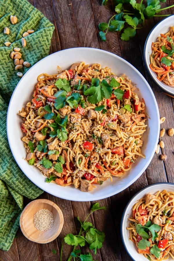 Peanut noodles with chicken in a large white bowl next to a small bowl of sesame seeds.