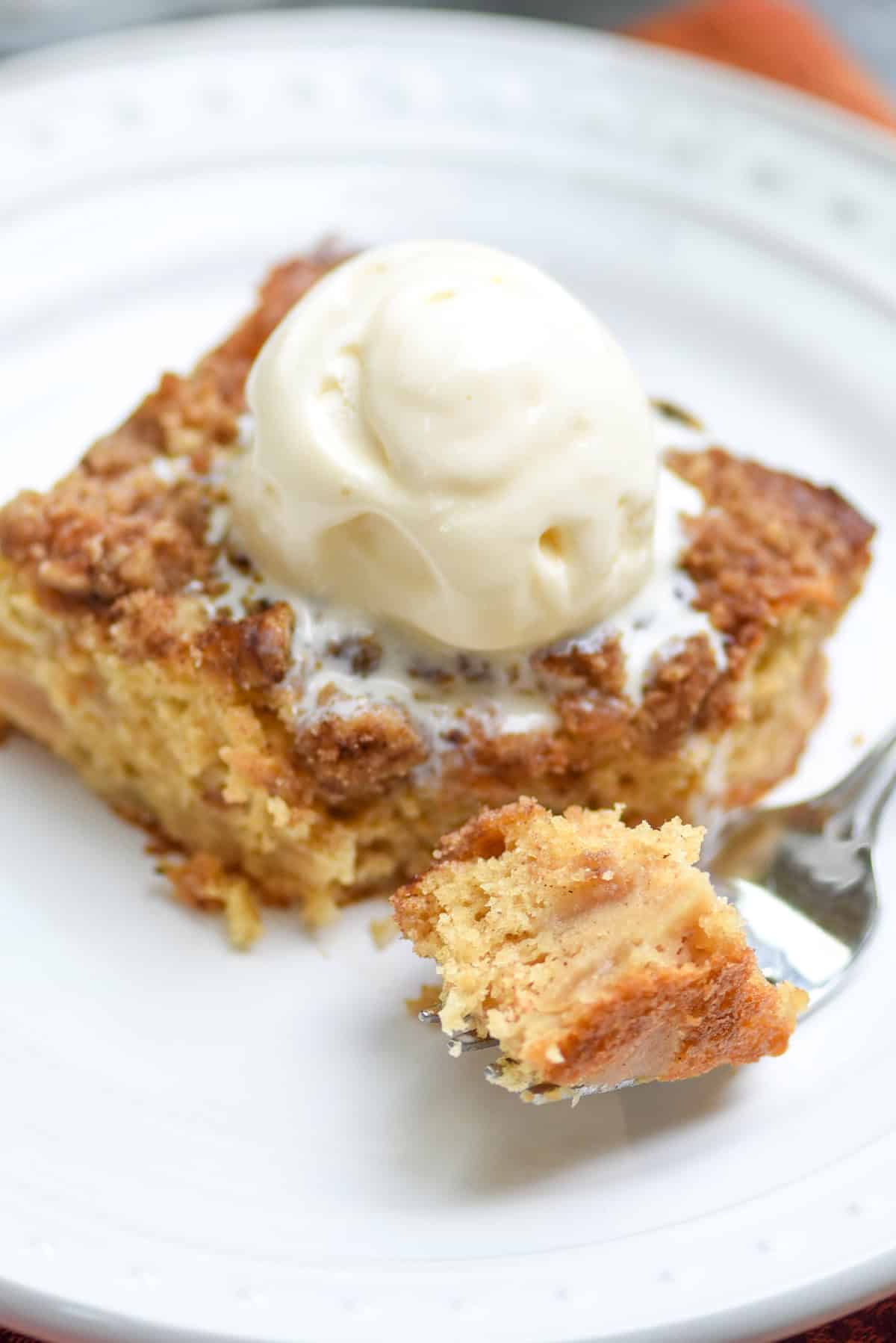 A forkwith a bite of cake resting on a plate with a slice of apple crumble cake topped with vanilla ice cream.