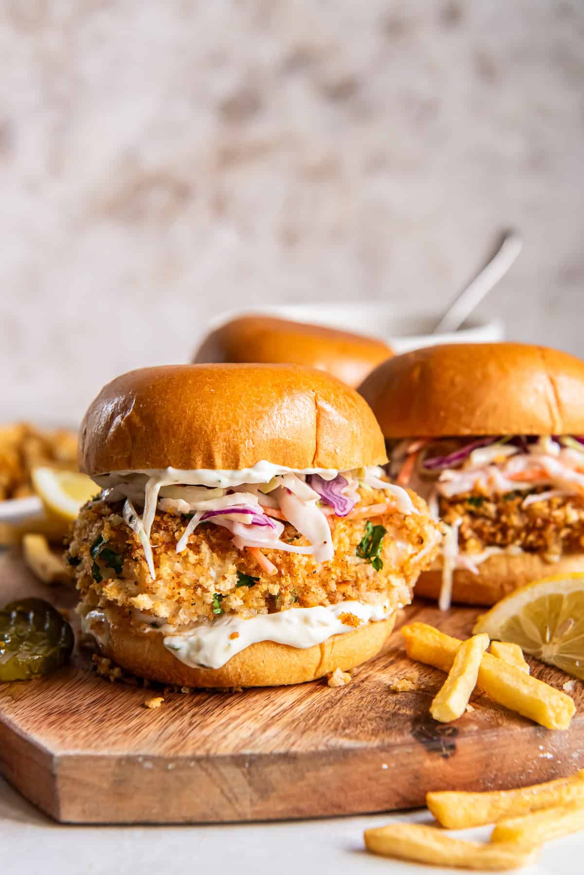 Three fish sandwiches with coleslaw on a wood board with French fries lying around it.