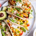 Plated fish stick tacos garnished with fresh toppings and surrounded by small bowls of salsa, sour cream, and shredded cheese.