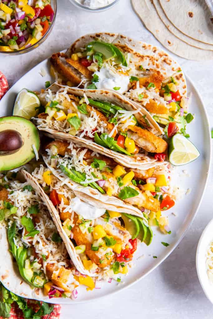 Plated fish stick tacos garnished with fresh toppings and surrounded by small bowls of salsa, sour cream, and shredded cheese.