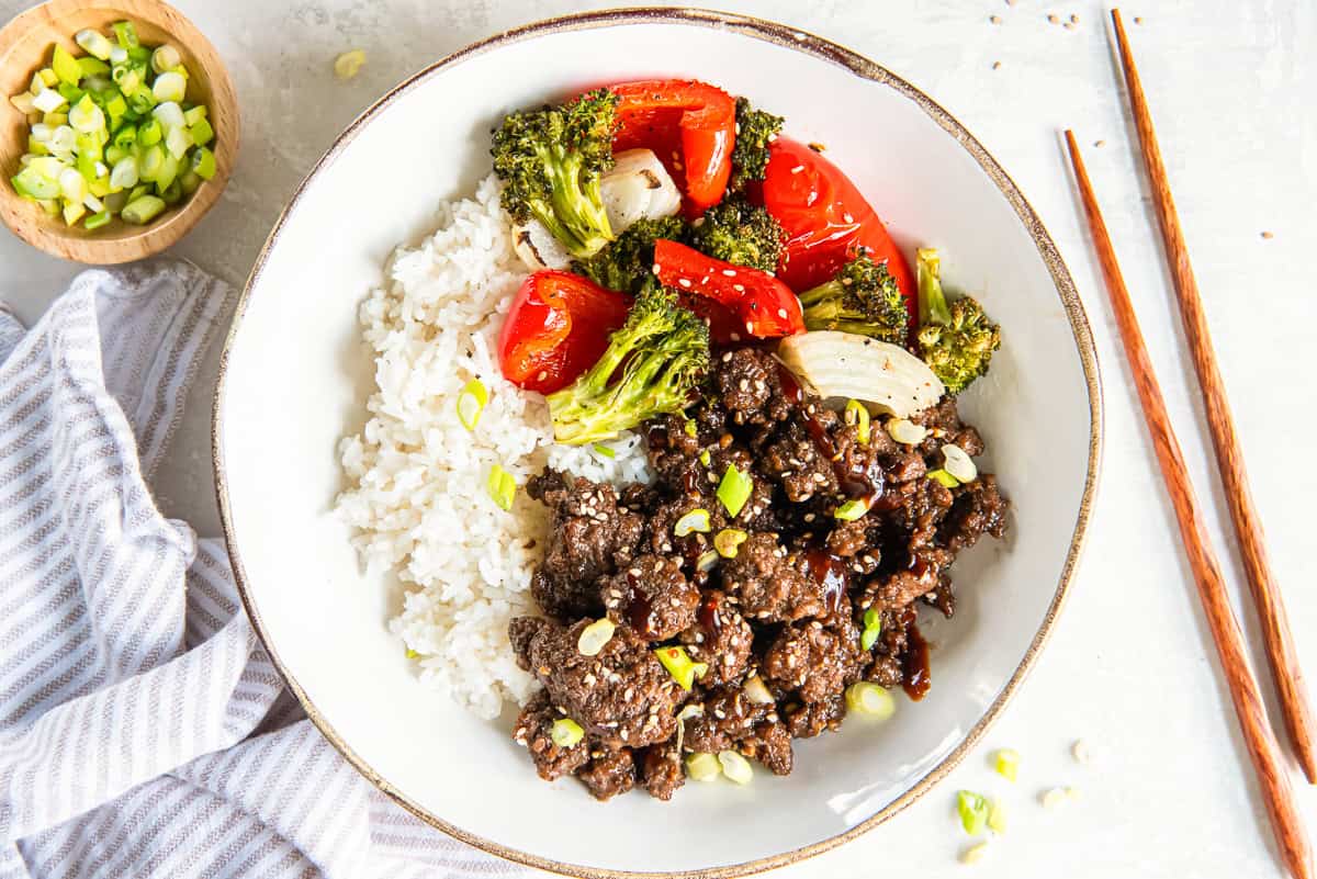 Chopsticks next to a white bowl filled with ground Korean beef, white rice and roasted vegetables.