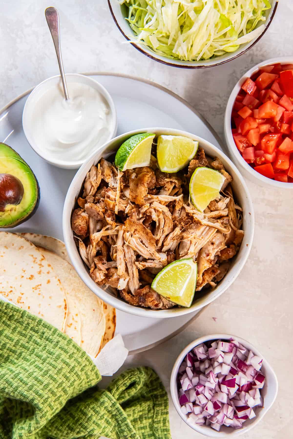 Mexican shredded chicken in a bowl surrounded by tortillas, sour cream, and other taco toppings.