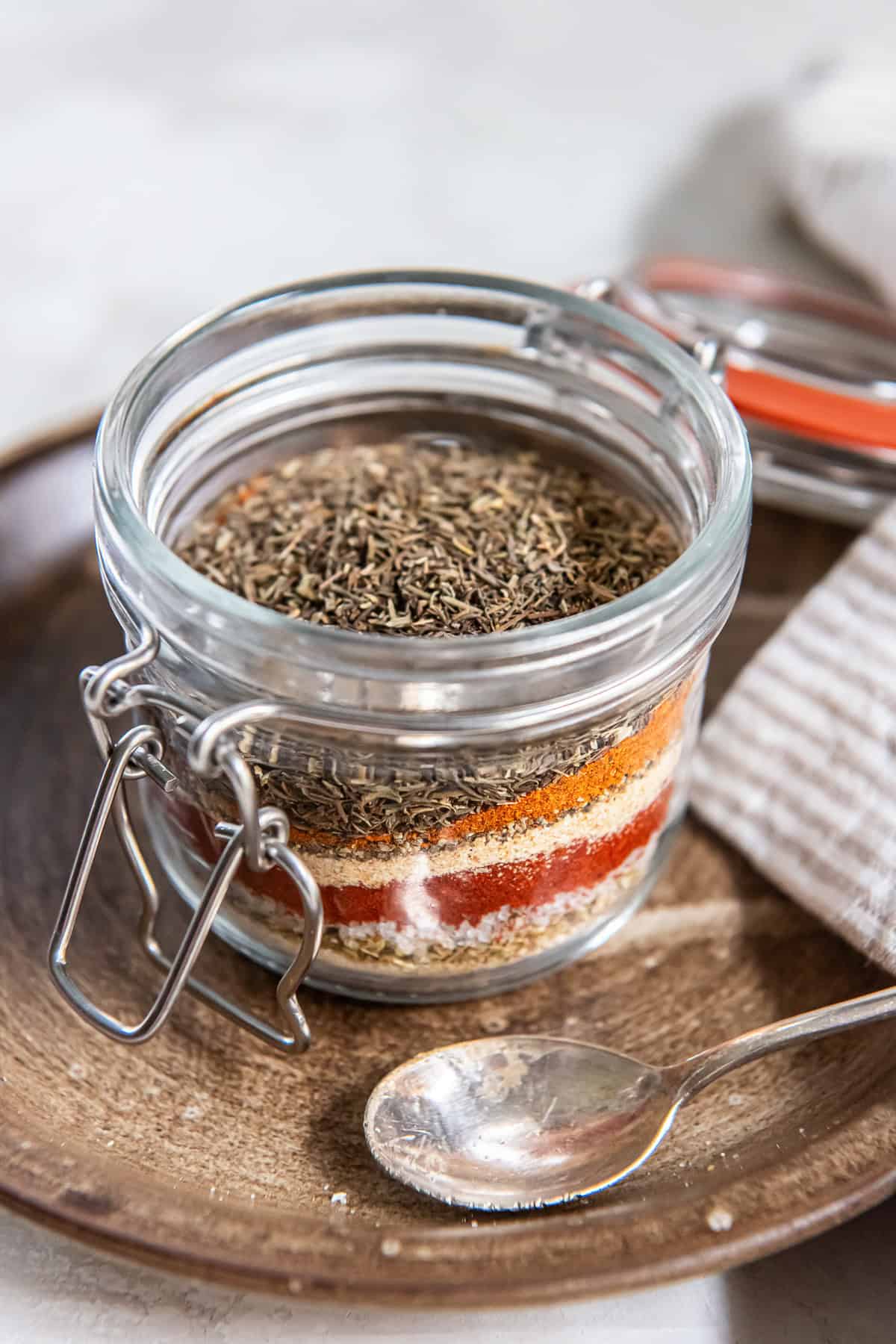 Glass jar of Cajun seasoning mix showing layers of dried herbs and spices with a silver spoon beside it.