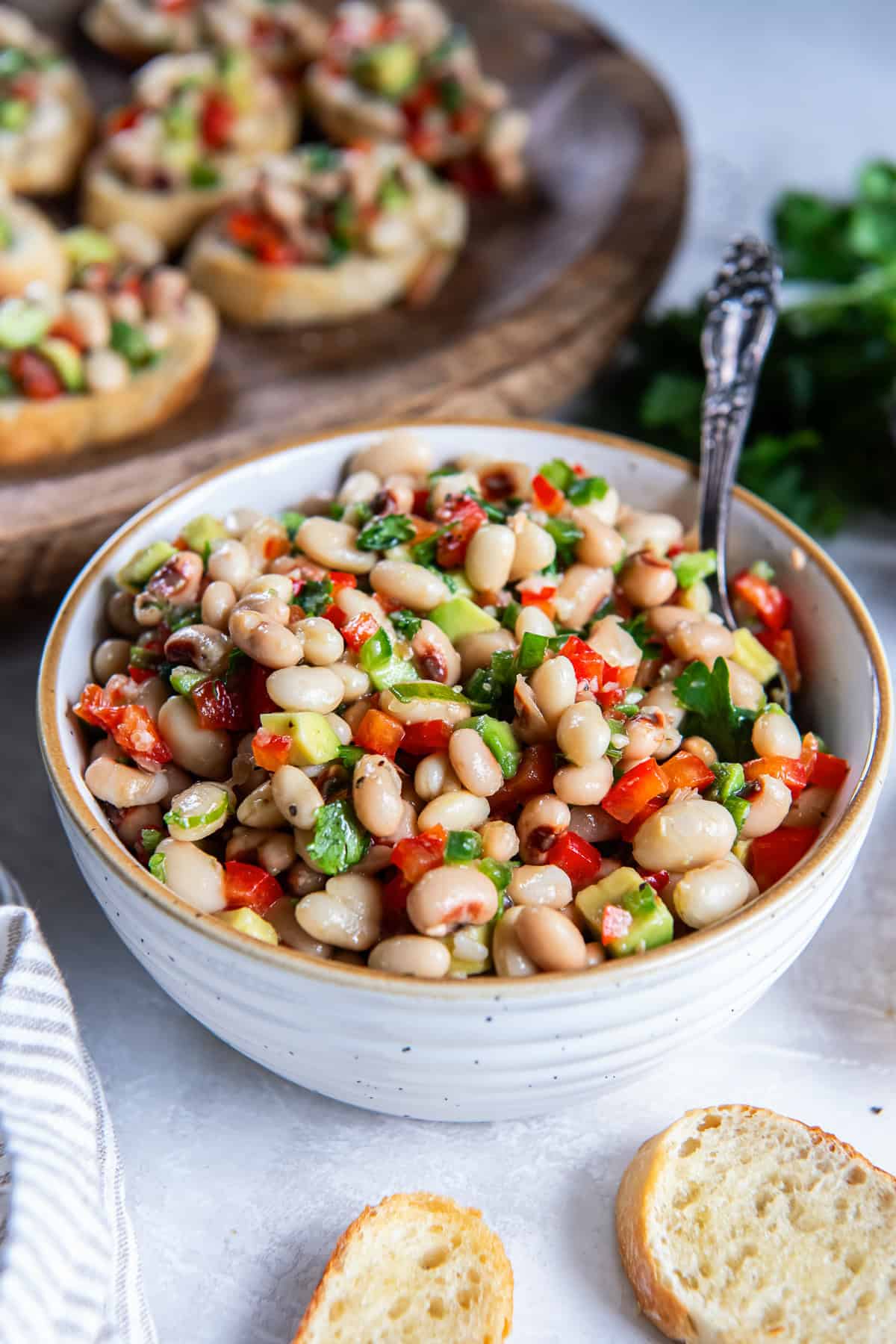 Bowl of white bean and black-eyed pea mixture with avocado, red bell pepper, jalapeño, and cilantro, with toasted baguette slices and crostini in the background.