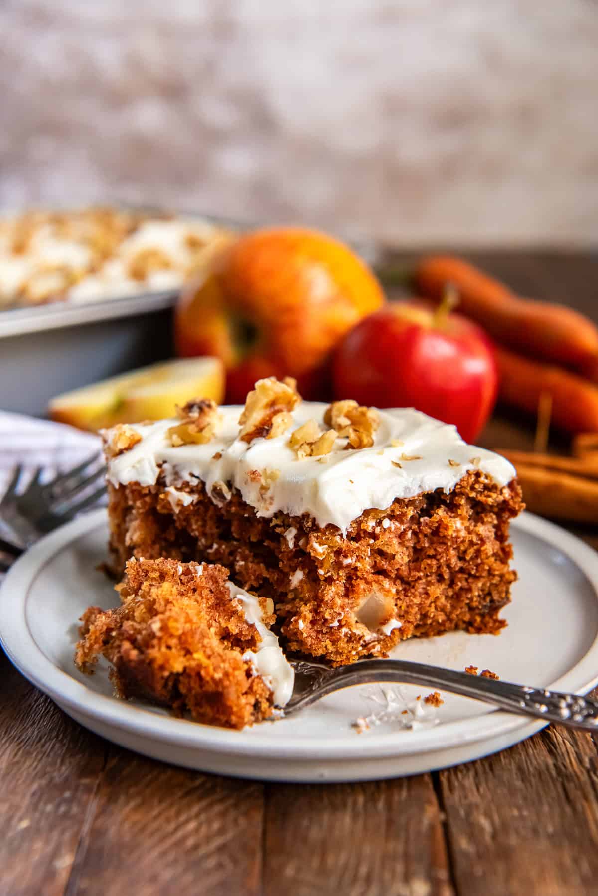 A fork resting on a plate with a slice of apple carrot cake with cream cheese frosting and walnuts.