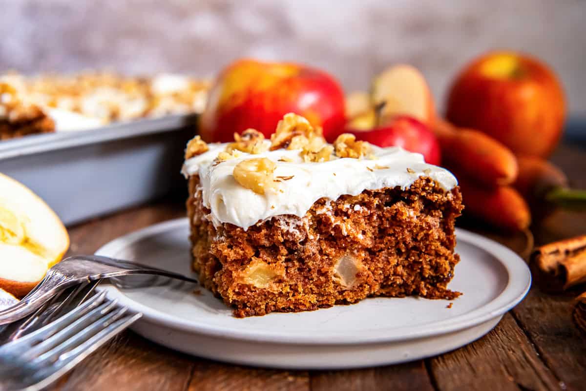 A slice of apple carrot cake on a white plate with apples in the background.