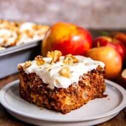A slice of apple carrot cake on a white plate with apples in the background.