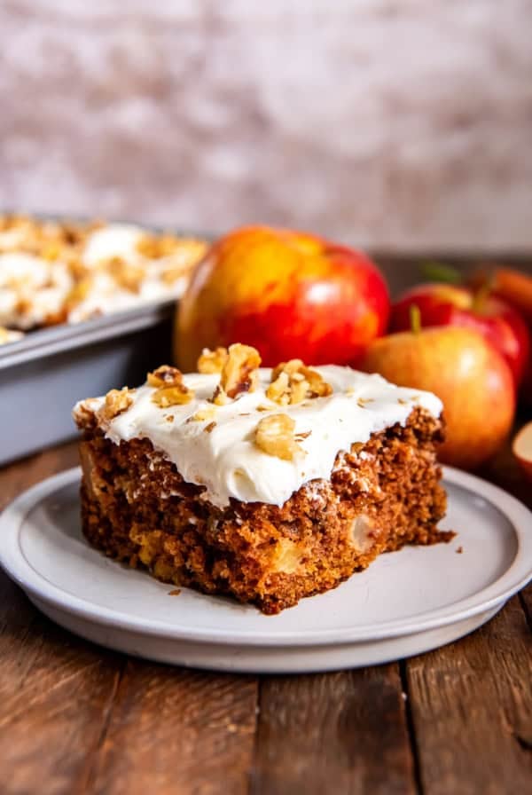 A slice of apple carrot cake on a white plate with apples in the background.