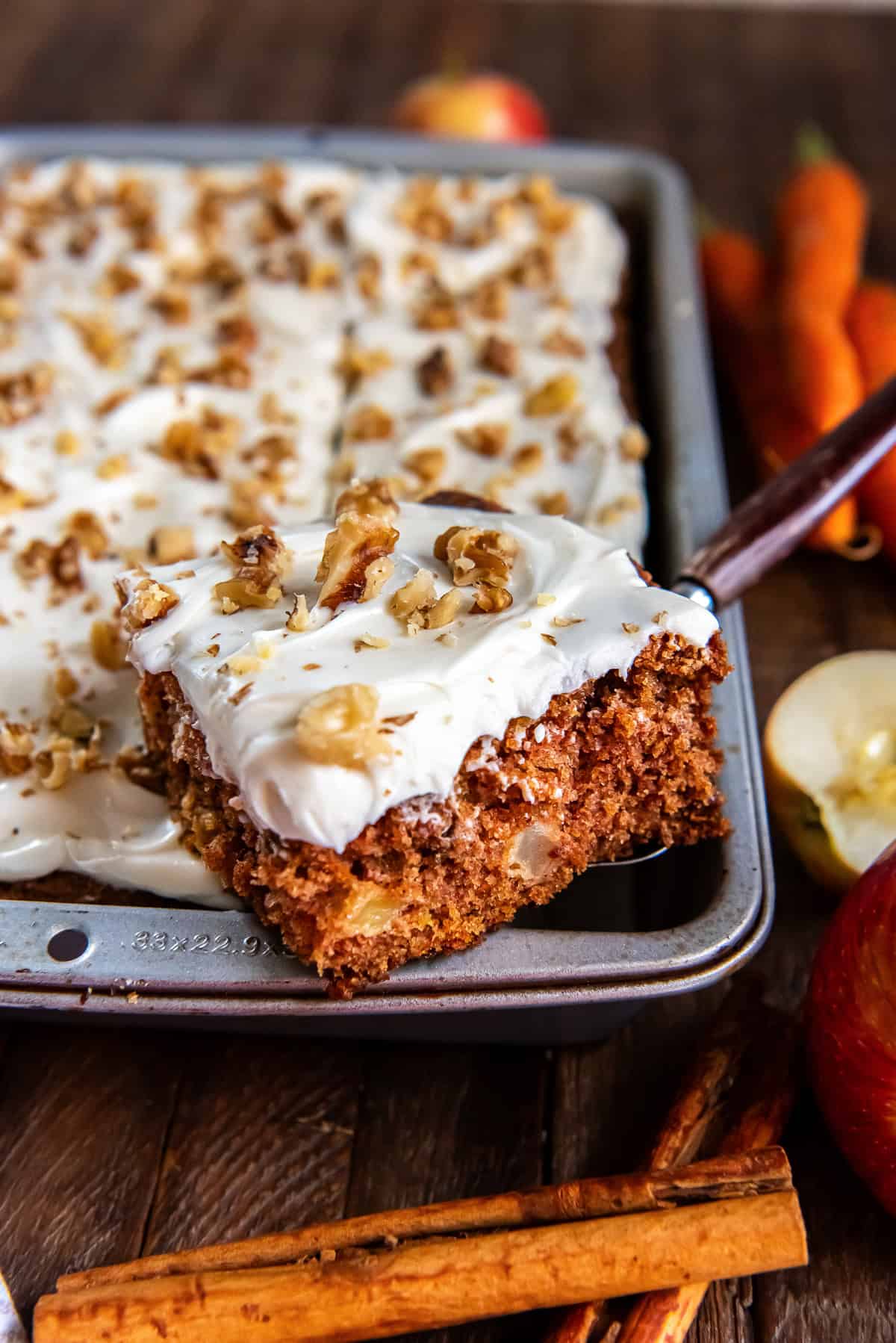 A spatula lifting a slice of apple carrot cake with cream cheese frosting from a baking pan.