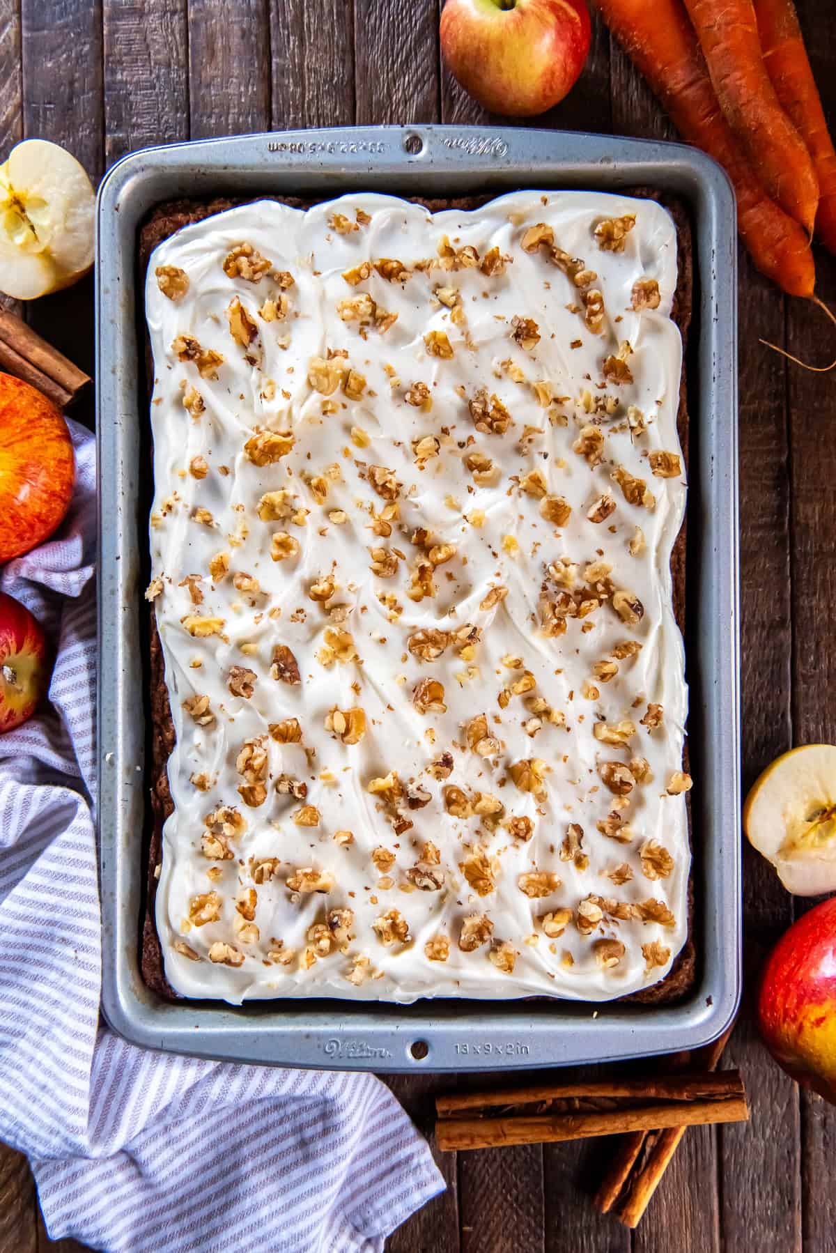 An apple carrot cake topped with cream cheese frosting and walnuts in a metal baking pan surrounded by appes and cinnamon sticks.