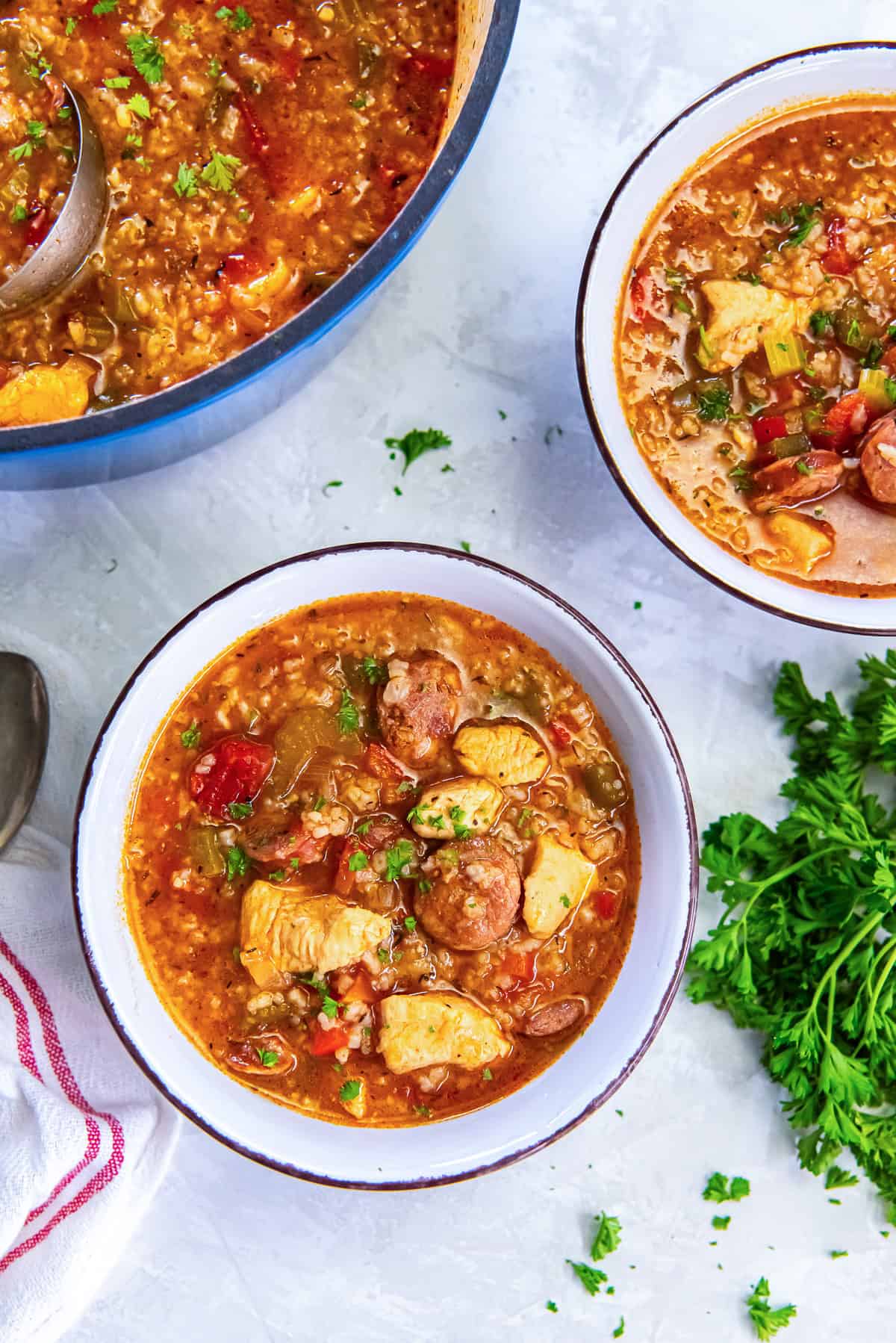 A blue Dutch oven of Jambalaya Soup with two bowls ready to serve and fresh parsley on the side.