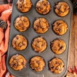 Pumpkin chocolate chip muffins in a muffin pan surrounded by fall leaves and an orange apron.
