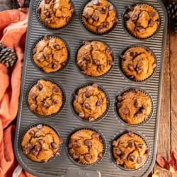 Pumpkin chocolate chip muffins in a muffin pan surrounded by fall leaves and an orange apron.