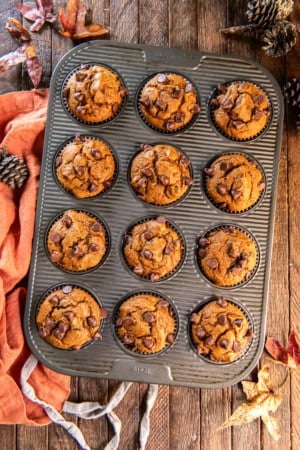 Pumpkin chocolate chip muffins in a muffin pan surrounded by fall leaves and an orange apron.