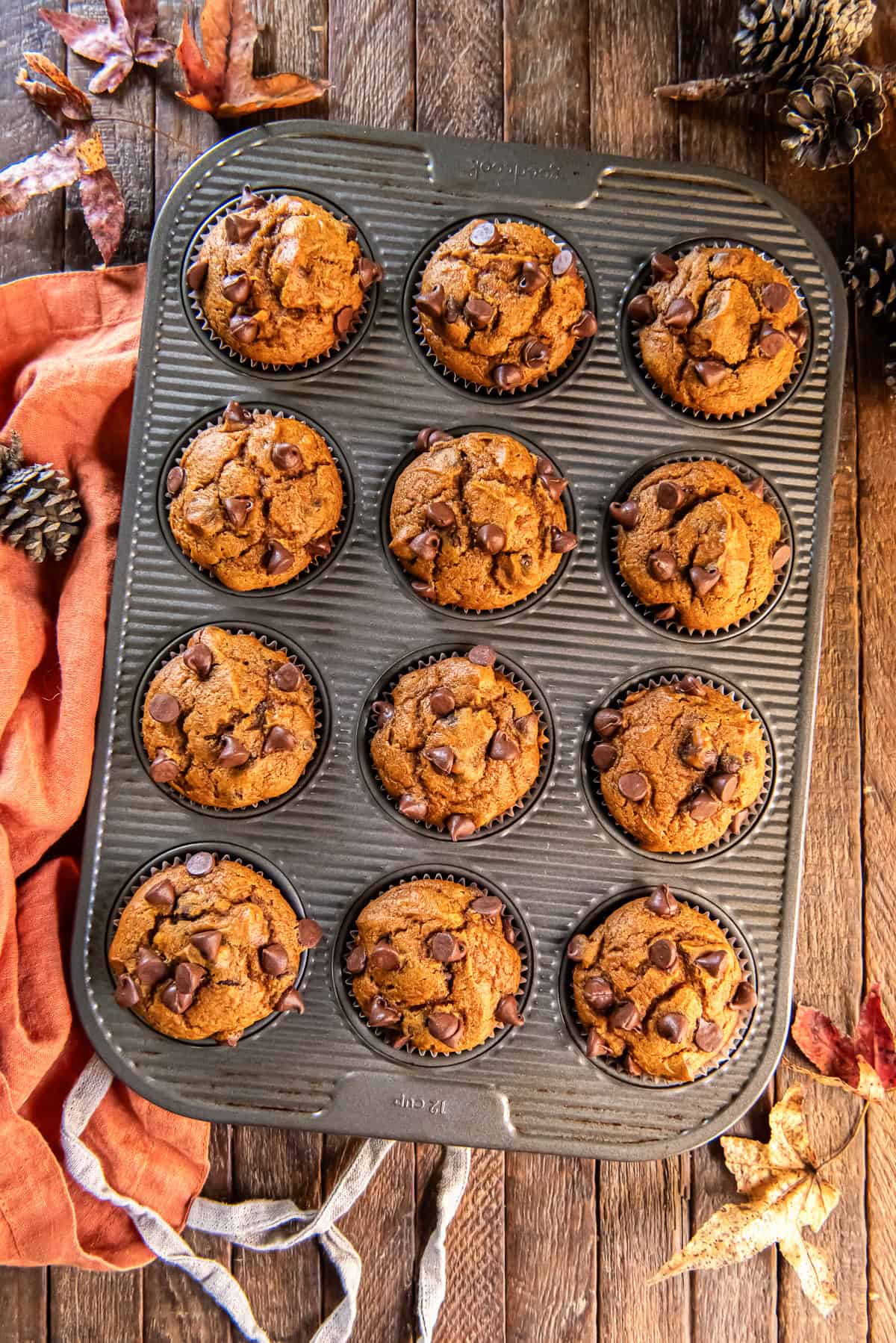 Pumpkin chocolate chip muffins in a muffin pan surrounded by fall leaves and an orange apron.