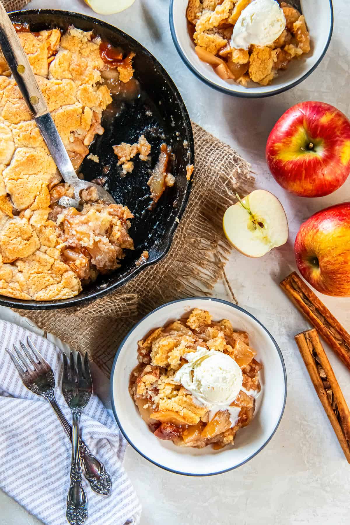 Bowl of warm skillet apple cobbler topped with melting vanilla ice cream, with a cast-iron skillet, apples, and cinnamon sticks in the background.