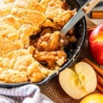 Skillet apple cobbler after a scoop, showing gooey cinnamon-apple filling and golden biscuit topping with a spoon.