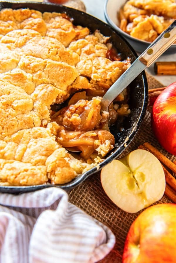 Skillet apple cobbler after a scoop, showing gooey cinnamon-apple filling and golden biscuit topping with a spoon.