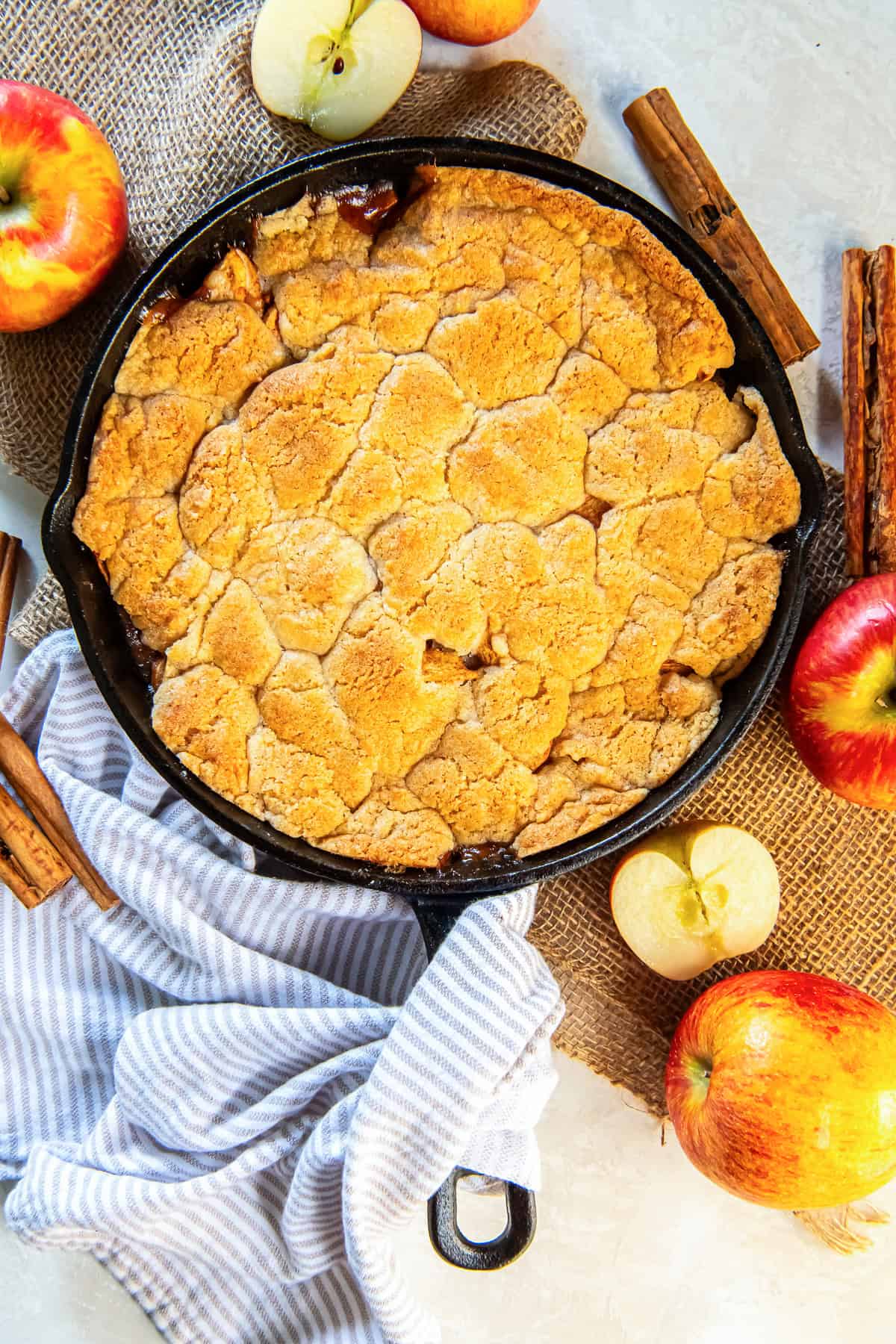 Apple cobbler in a cast iron skillet with a kitchen towel wrapped around the handle and surrounded by apples and cinnamon sticks.