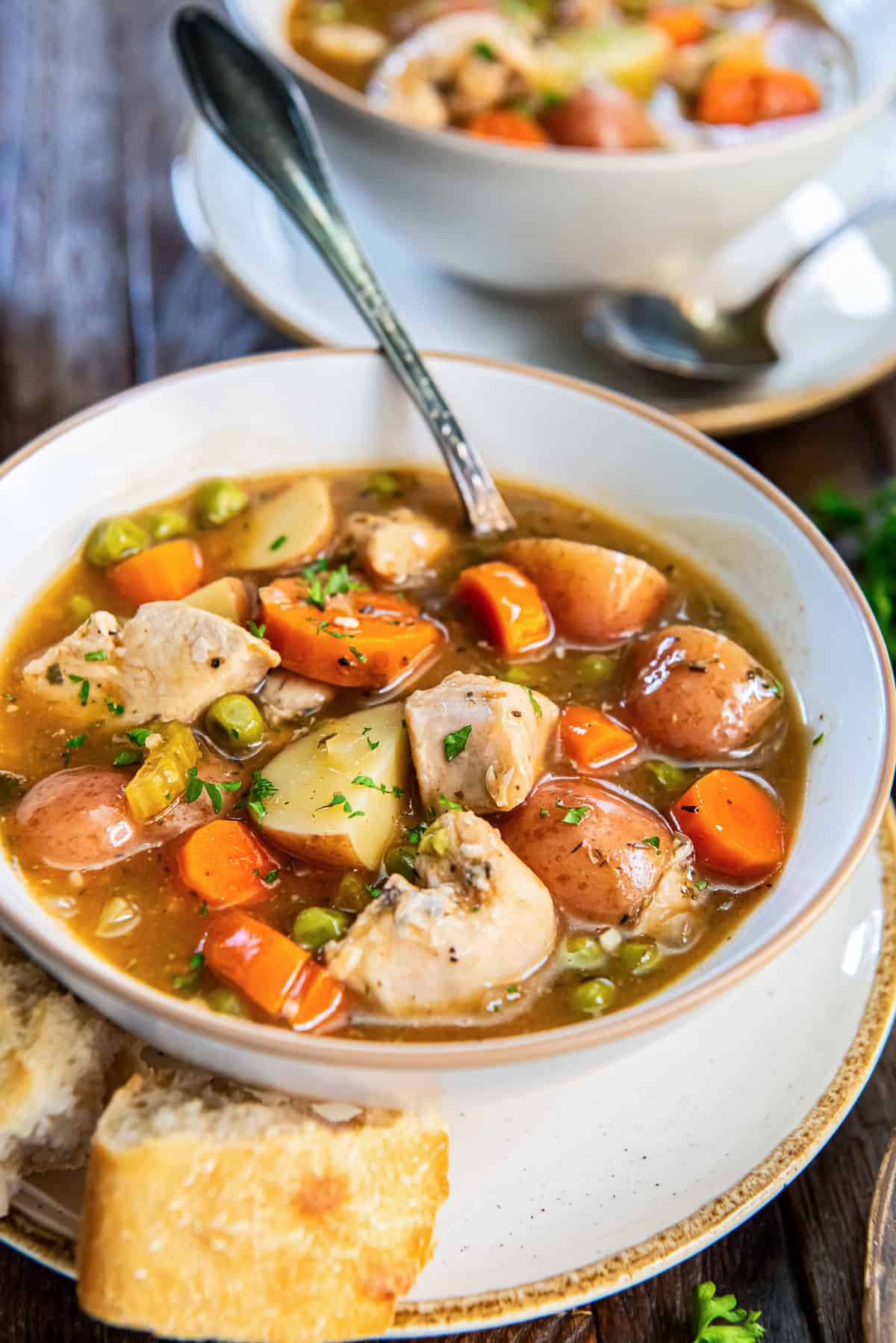Bowl of slow cooker chicken stew with carrots, baby red potatoes, peas, and parsley; a second bowl blurred in the background.