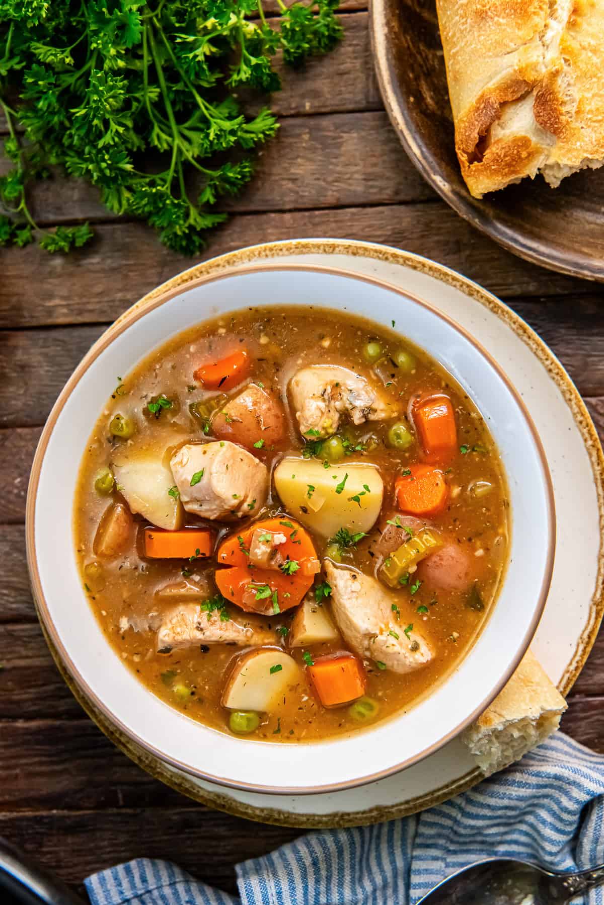 Chicken stew with carrots, red potatoes, peas, and parsley in a white bowl on a rustic table with bread.
