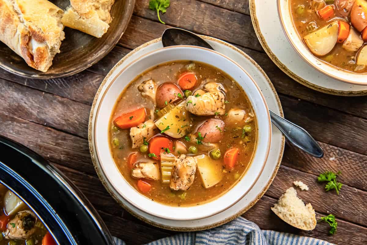 Bowl of slow cooker chicken stew with carrots, baby red potatoes, and peas, with crusty bread on the side.