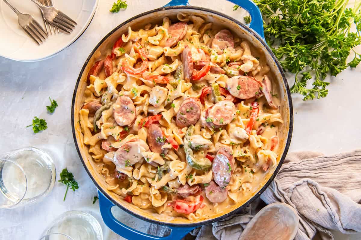 A blue pan filled with creamy Cajun Chicken and Sausage Pasta with bell peppers, parsley, and glasses of white wine nearby.