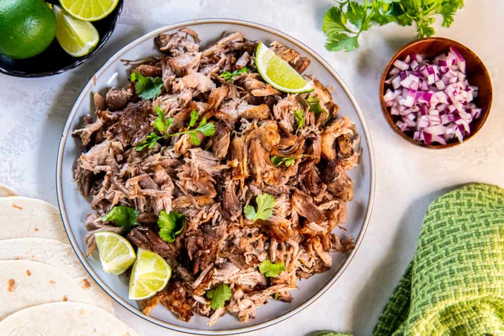 Shredded pork carnitas on round platter with lime wedges next to a small bowl of diced red onion.