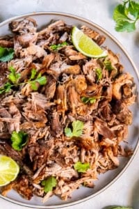 Shredded pork carnitas on round platter with lime wedges next to a small bowl of diced red onion.