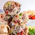 Two carnitas burritos cut in half and stacked on a wooden board, showing layers of shredded pork, rice, beans, guacamole, and salsa with lime wedges and cilantro in the background.