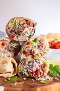Two carnitas burritos cut in half and stacked on a wooden board, showing layers of shredded pork, rice, beans, guacamole, and salsa with lime wedges and cilantro in the background.
