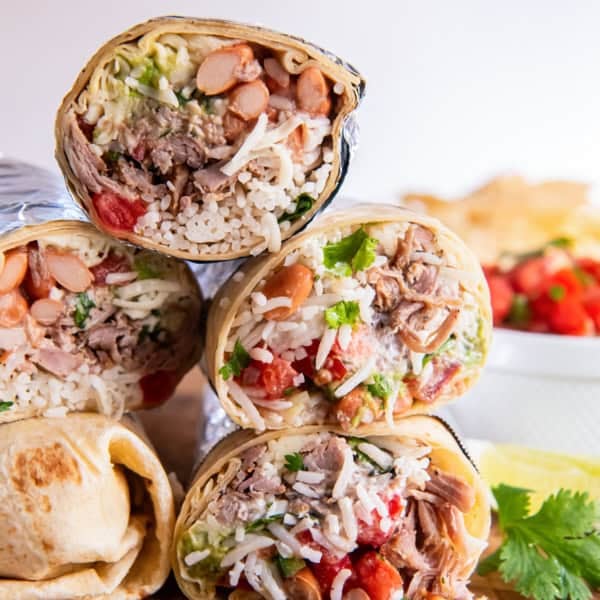 Two carnitas burritos cut in half and stacked on a wooden board, showing layers of shredded pork, rice, beans, guacamole, and salsa with lime wedges and cilantro in the background.