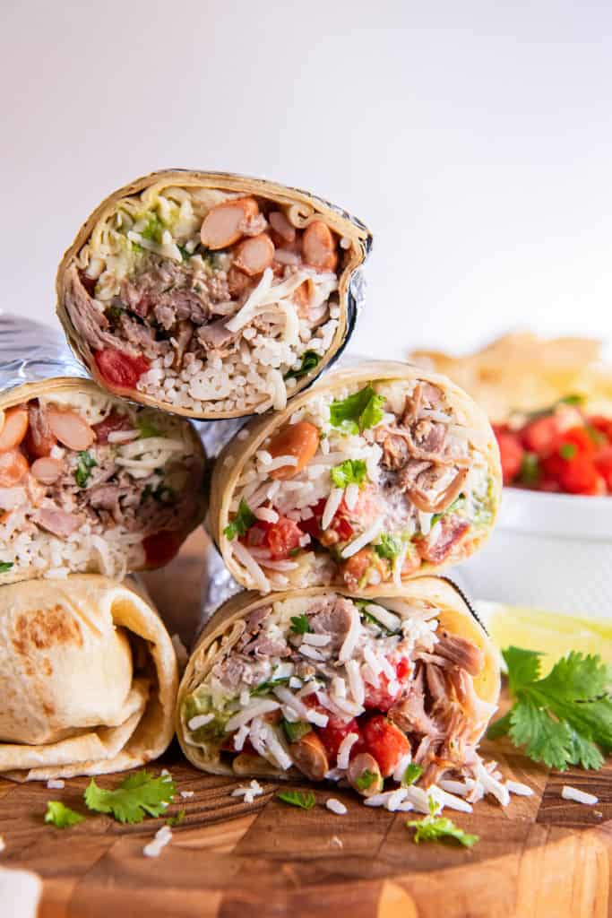 Two carnitas burritos cut in half and stacked on a wooden board, showing layers of shredded pork, rice, beans, guacamole, and salsa with lime wedges and cilantro in the background.