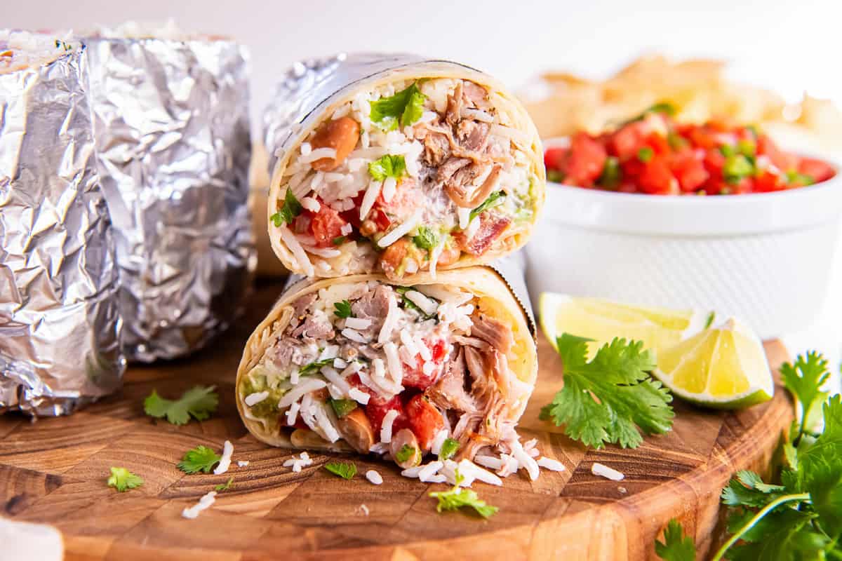 A carnitas burritos wrapped in foil and cut in half and stacked on a wooden board, showing layers of rice, shredded pork, beans, guacamole, and salsa.