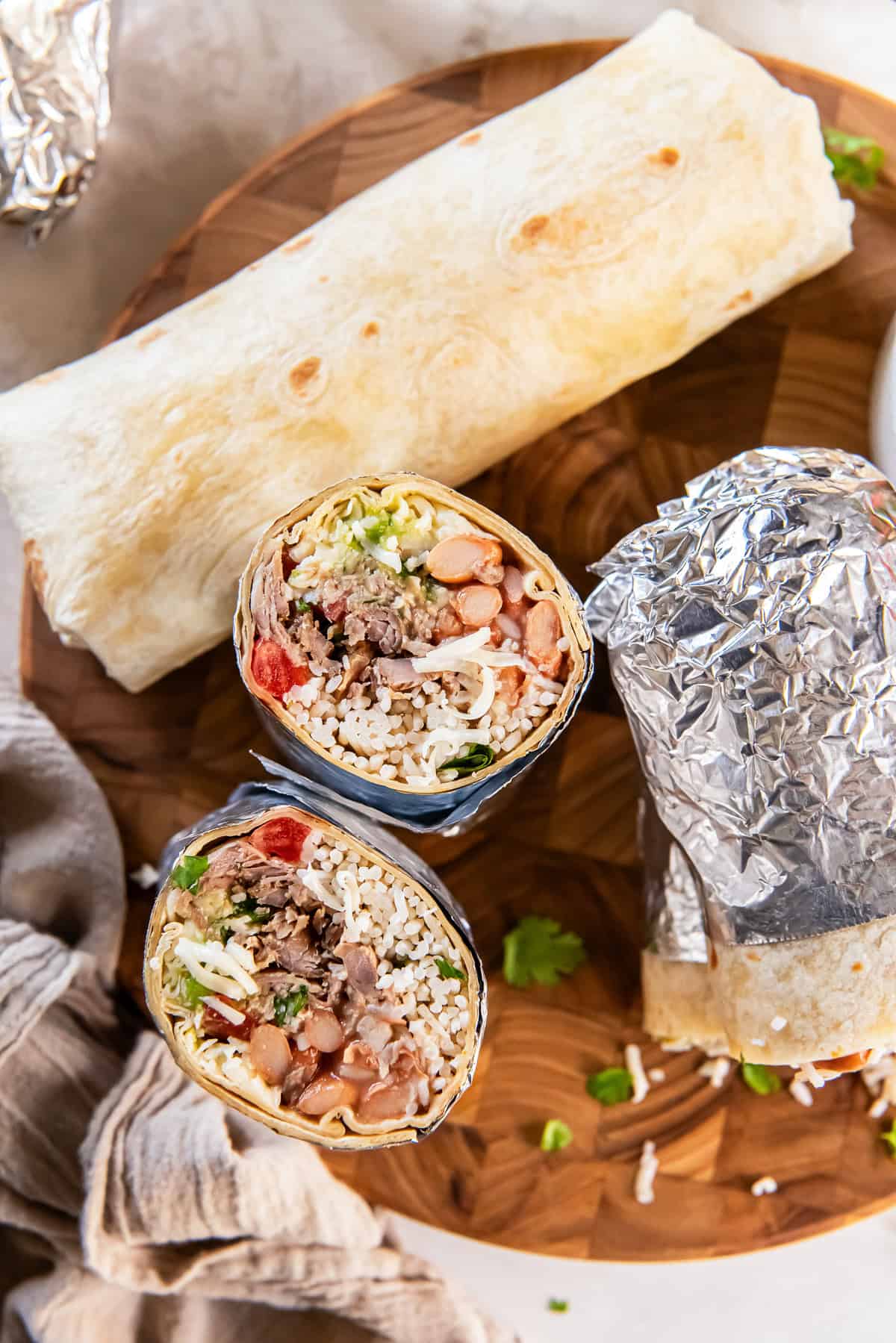Two carnitas burritos cut in half and stacked on a wooden board, showing rice, beans, shredded pork, guacamole, and pico de gallo with foil-wrapped burritos, chips, and lime wedges in the background.