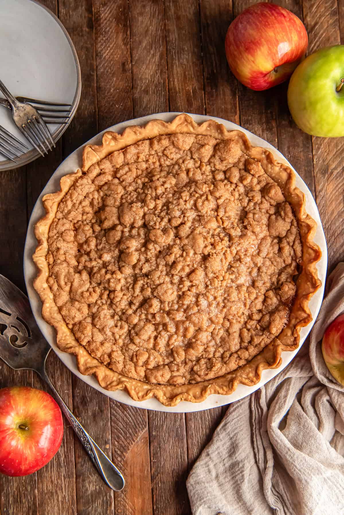 A whole Dutch apple pie with crumb topping on a dark wood surface with apples.