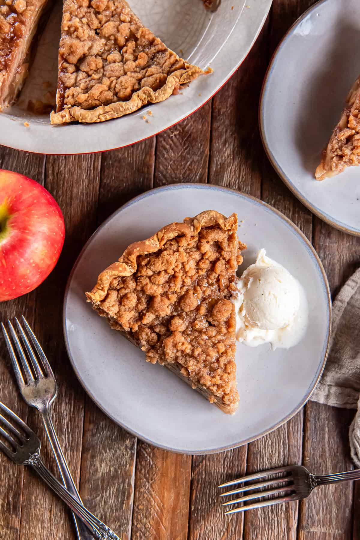 A slice of Dutch apple pie with a crumb topping served on a plate with a fork next to fresh apples and rustic wooden table background.