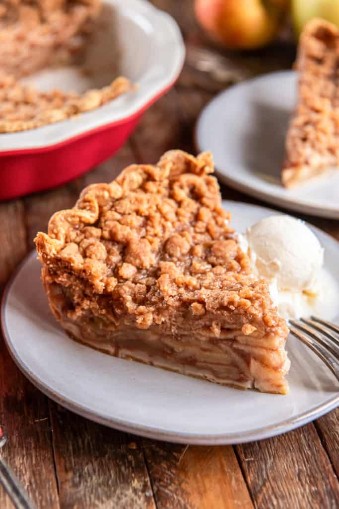 A slice of Dutch apple pie with a crumb topping served on a plate with a scoop of vanilla ice cream, with the remaining pie in the background.
