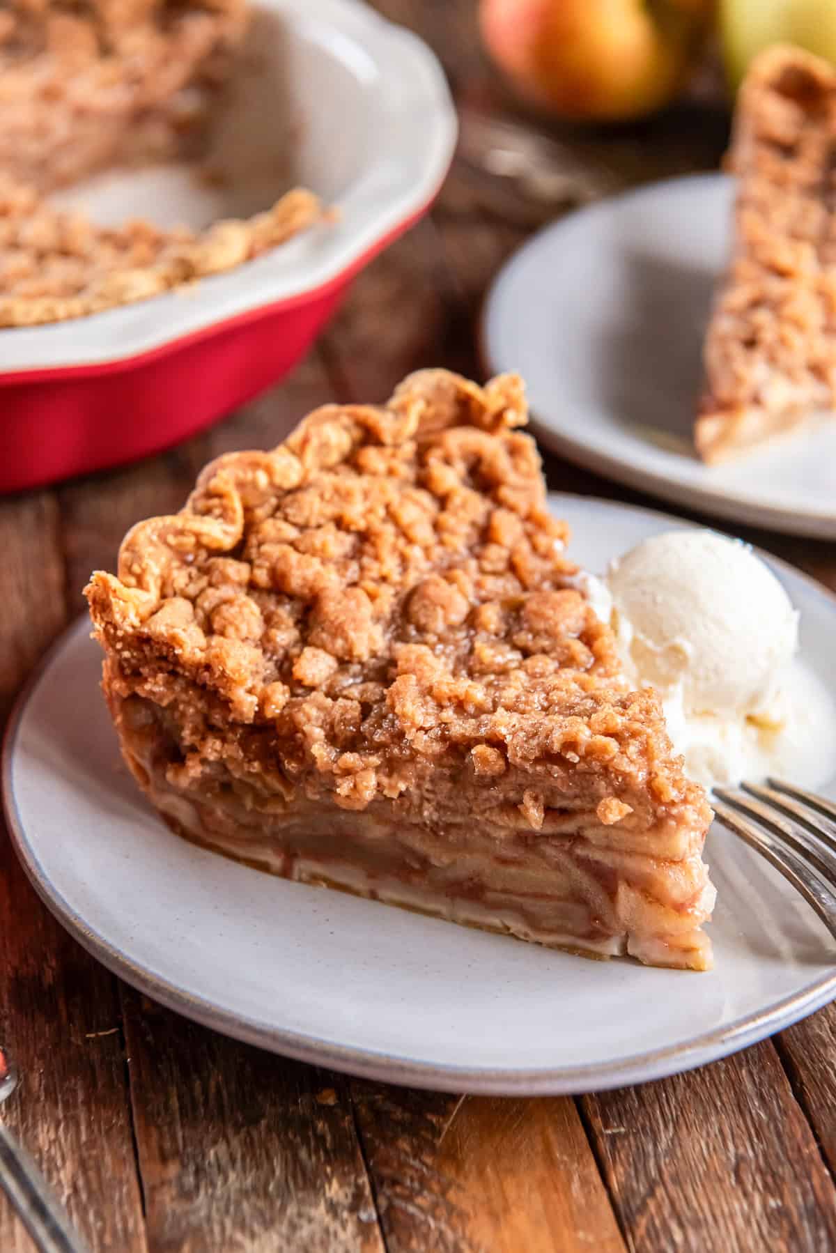 A slice of Dutch apple pie with a crumb topping served on a plate with a scoop of vanilla ice cream, with the remaining pie in the background.