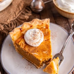 Slice of Hazelnut Pumpkin Pie topped with whipped cream and sprinkled with cinnamon, with a forkful ready to eat on a rustic plate.